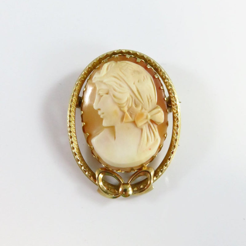 This vintage cameo brooch features a stunning profile of a woman, her hair adorned with a bow. The detailed carving is set in a 14k gold-filled prong setting. The outer edge of the oval frame boasts a lovely textured sawtooth design that gracefully curves into a bow at the bottom. This elegant cameo is in excellent condition and shows no signs of cracking