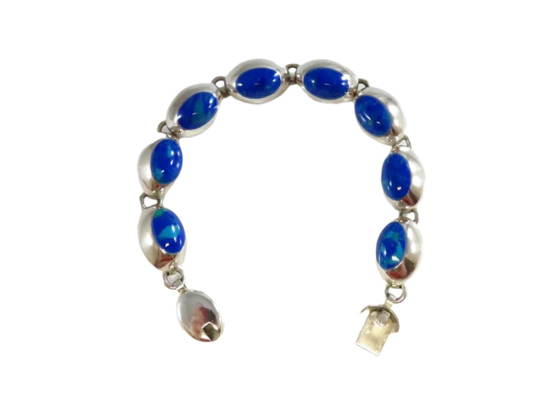 This substantial 7 ¾-inch Southwestern-style bracelet features eight oval, polished cabochon stones of unknown origin. Each stone measures approximately 9mm by 13mm and is bezel-set in an eye-shaped sterling silver setting, creating a playful, almost hypnotic effect. The bracelet secures with a slide lock clasp. Evoking the appearance of azurite, the “stone of heaven,” the piece showcases rich blue and green tones. Elegantly crafted in sterling silver.